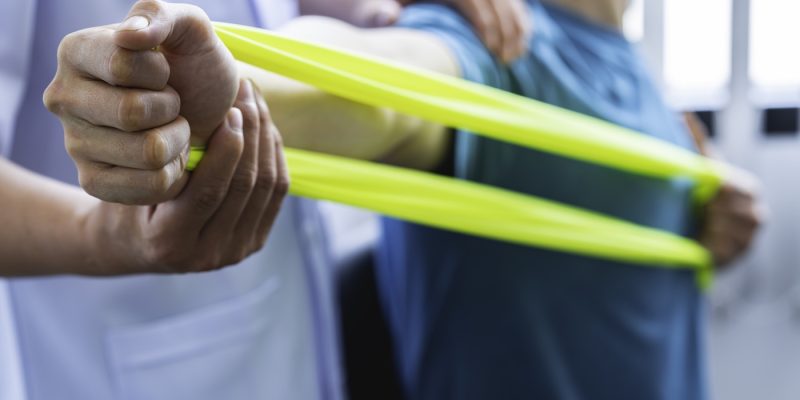 Physical therapist gives resistance band exercises. About the arms and shoulders of male office workers, office syndrome, male patient. Physiotherapy concept.